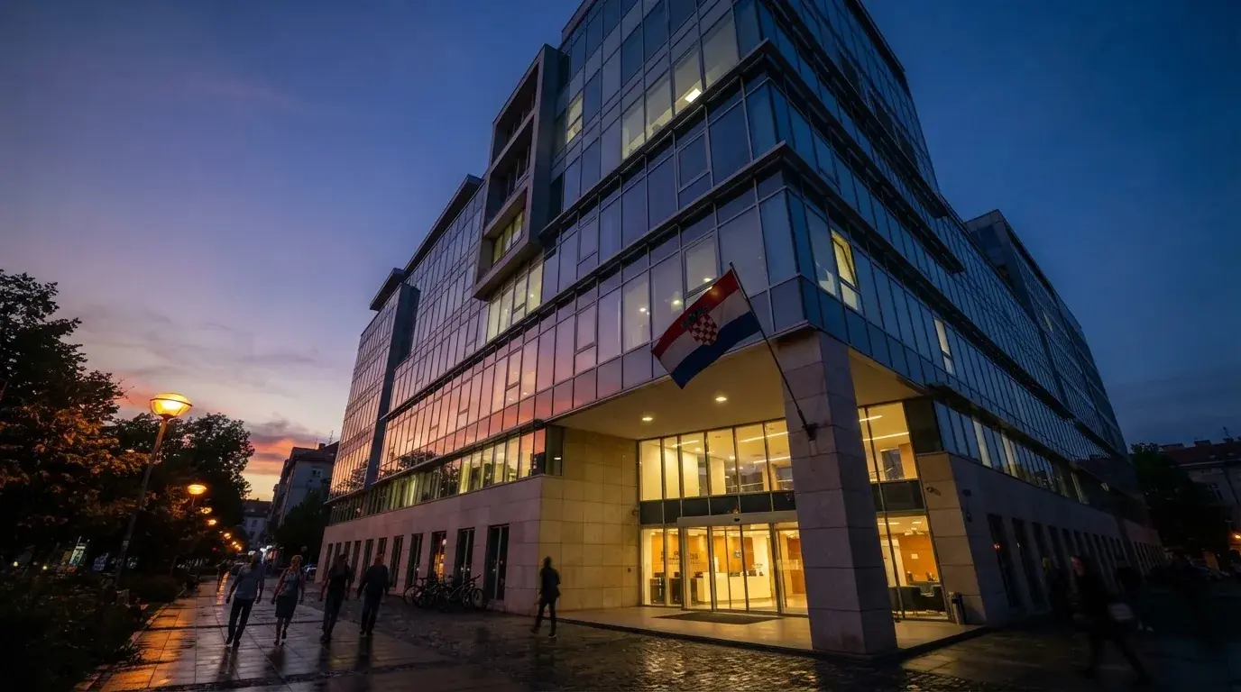 Fasada moderne državne zgrade u Zagrebu s ravnim staklenim pročeljem i hrvatskom zastavom, snimljena u sumrak.